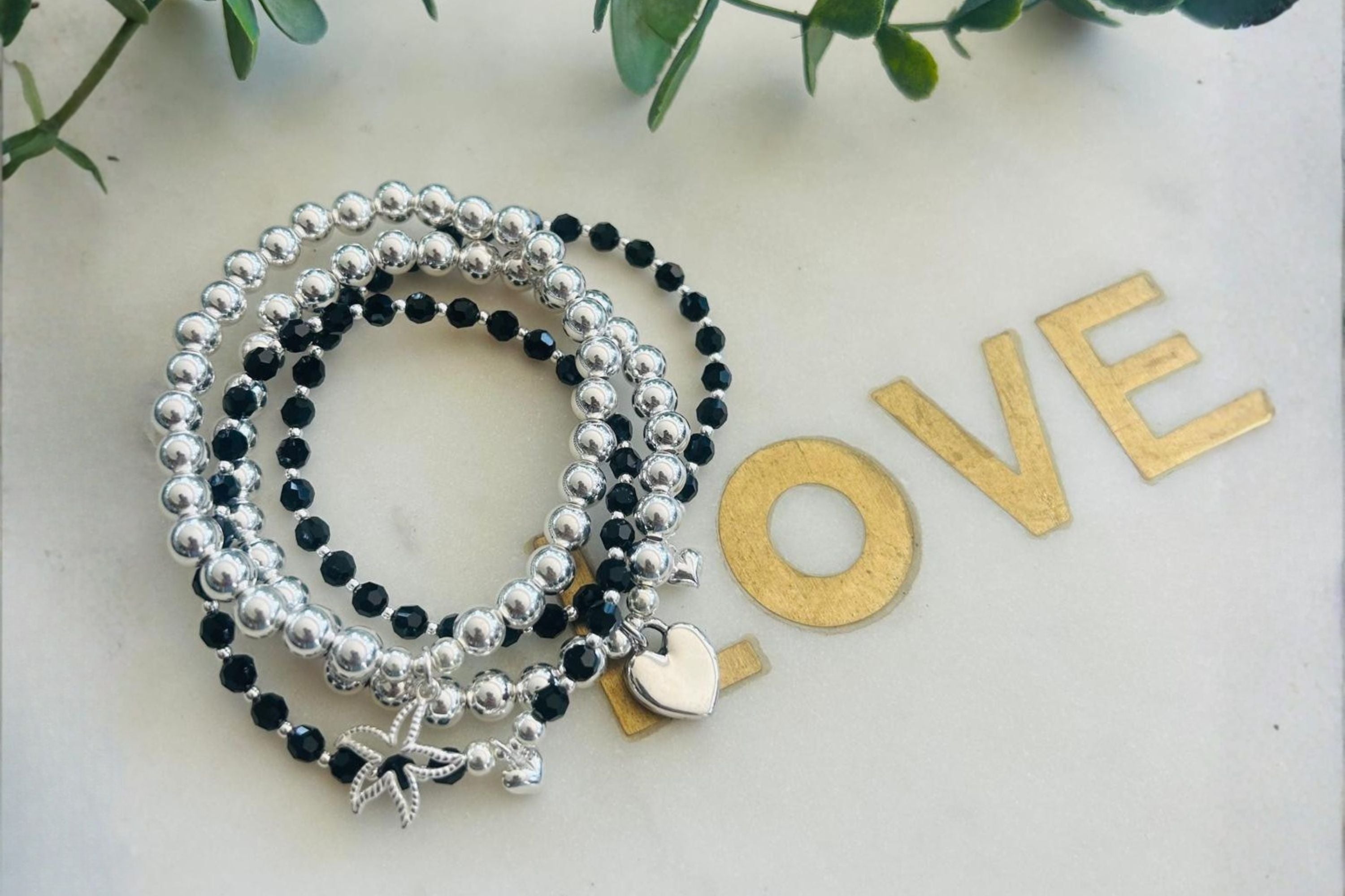 Three beaded stretch bracelets with silver and black beads, featuring a silver star charm, a silver heart charm, and a small rhinestone star charm, arranged on a light surface with gold 'LOVE' letters and green leaves in the background.